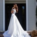 A woman in a white, off-the-shoulder wedding gown, reminiscent of stunning formal dresses, stands on stairs outdoors, smiling and touching her lips with one hand. Her dress has a long, flowing train. She is positioned in front of a dark-paneled doorway with a white decorative arch. Angels Formal Wear