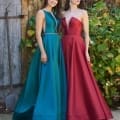 Two women stand side by side in front of a rustic wooden door with greenery and flowers in the background. They are both wearing elegant, floor-length designer bridesmaid dresses; one in emerald green with a beaded belt, and the other in deep red with a plunging neckline. Angels Formal Wear