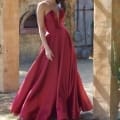 A woman stands outdoors wearing a strapless red gown with a full skirt and deep neckline. She is under an archway, with a rustic wall and closed wooden door in the background. The sunlight creates soft shadows, enhancing the elegance of her formal dress and pose. Angels Formal Wear