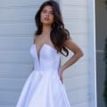 A woman poses in a strapless white wedding gown with a deep V-neckline, reminiscent of designer evening wear. She stands against a light-colored paneled wall, looking slightly to the side. Her long, dark hair is styled in soft waves, and her left hand rests on her hip as her right hand is in the dress pocket. Angels Formal Wear