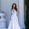 A woman stands outside beside a building, wearing a strapless white designer evening wear gown with a deep V-neckline and a flowing skirt. Her left hand rests in her pocket as she gazes forward. The background includes a white decorative planter and some greenery. Angels Formal Wear