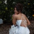 A woman in the GRACE TC322 white backless dress, featuring an intricate crisscross tie design, kneels on one knee next to an orange tree while holding white flowers. She gazes over her shoulder, showcasing her elegant updo hairstyle in an outdoor garden setting—an exquisite vision that could inspire many seeking wedding dresses online in Australia. Angels Formal Wear