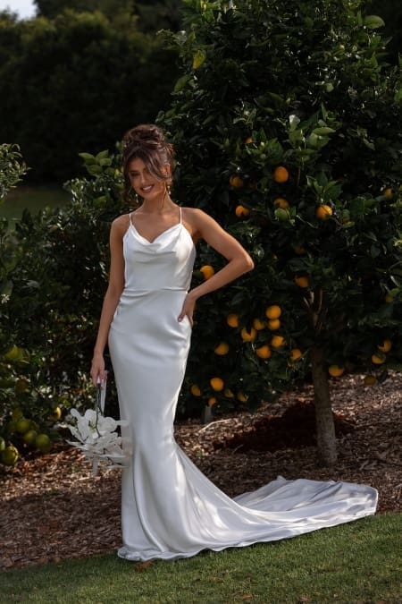 A woman in a sleeveless white satin GRACE TC322 bridal gown, purchased from Wedding Dresses Online Australia, stands outdoors beside an orange tree. She holds a white flower bouquet and has her other hand on her hip. The lush green background and fallen leaves enhance the scene's natural beauty. Angels Formal Wear