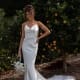 A woman in a sleeveless white satin GRACE TC322 bridal gown, purchased from Wedding Dresses Online Australia, stands outdoors beside an orange tree. She holds a white flower bouquet and has her other hand on her hip. The lush green background and fallen leaves enhance the scene's natural beauty. Angels Formal Wear