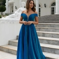 A woman stands on outdoor steps in front of a white building, wearing a flowing, off-shoulder teal gown from BETH PO861S. The dress features a deep V-neck and a fitted bodice. A white statue of a reclining figure is visible in the background. She is looking to the left.
 Angels Formal Wear