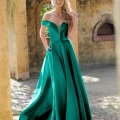 A woman stands outdoors in front of a rustic wall, wearing an elegant emerald green off-the-shoulder gown from BETH PO861S. The dress features a fitted bodice and flows into a full, floor-length skirt. She smiles as she lightly touches her hair, which falls in loose waves over one shoulder. Angels Formal Wear