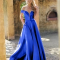 A woman in a vibrant blue, off-the-shoulder BETH PO861S gown stands outdoors against a rustic wall, smiling and gently touching her hair. Sunlight highlights her flowing dress and casts shadows on the cobblestone path beneath her. Angels Formal Wear