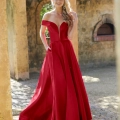 A person stands outdoors in front of a warm-toned, textured wall and archway. Draped in a vibrant, floor-length red gown from BETH PO861S, with an off-the-shoulder neckline and pleated skirt, their hair is styled in loose waves, and they have a pleasant expression. Angels Formal Wear