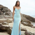 A woman stands on a rocky beach wearing a silky, light blue, floor-length dress with a thigh-high slit from PIPER PO858. She has long brown hair and is looking directly at the camera. The ocean and a cloudy sky are visible in the background. Angels Formal Wear