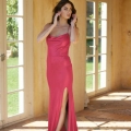 A woman stands barefoot in an elegant hallway adorned with large glass doors and a tiled floor. She is wearing a form-fitting, red satin PIPER PO858 gown with thin straps and a thigh-high slit, and she is adjusting one of the straps with a thoughtful expression. Angels Formal Wear