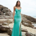 A woman stands on a rocky beach wearing PIPER PO858 from Dress Shop Sunshine Coast, featuring a cowl neckline and a high slit. Her long hair is down, and she gazes at the camera. The sky is overcast, and waves are visible in the background. Angels Formal Wear