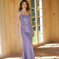 A woman stands indoors on a tiled floor, wearing a silky, floor-length lavender dress with thin straps. She has long, wavy hair and neutral makeup. Behind her is a large window framed by a light, textured wall. Sunlight streams in from the PIPER PO858, illuminating the space. Angels Formal Wear