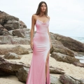 A woman stands on a rocky beach wearing a long, silky pink PIPER PO858 from Dress Shop Sunshine Coast, with thin straps and a thigh-high slit. She has long, straight hair and a serious expression. The background features large rocks and the ocean under a cloudy sky. Angels Formal Wear