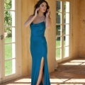 A woman wearing a teal, floor-length, sleeveless PIPER PO858 dress with a thigh-high slit stands in a sunlit room with cream-colored walls and large windows. She is adjusting her hair with both hands while looking off to the side. Barefoot, she seems ready to step into the Dress Shop Sunshine Coast. Angels Formal Wear