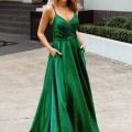 A woman wearing a long, emerald-green satin MONROE PO891 stands outdoors on a paved area. The dress features thin spaghetti straps, a V-neckline, and pockets. She holds her hands in the pockets and looks confidently at the camera. A building and greenery are visible in the background of this Dress Shop Sunshine Coast photo. Angels Formal Wear