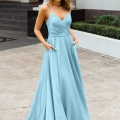 A woman stands outside on a paved area, wearing a light blue, sleeveless, floor-length gown with thin straps and a fitted bodice. She has her hands in the dress pockets and is looking at the camera. Behind her is a building with a large lion statue, reminiscent of the styles found in MONROE PO891. Angels Formal Wear