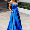 A woman stands confidently outdoors, wearing a vibrant blue MONROE PO891 with thin straps and a V-neckline. The dress features a fitted bodice with a slight drape and a flowing, floor-length skirt. She has her hands in the dress pockets and is posing in front of Dress Shop Sunshine Coast. Angels Formal Wear