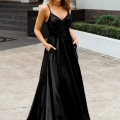 A woman is wearing a black, floor-length, sleeveless gown with thin straps and a V-neckline. She stands outdoors on a concrete pathway, posing with her hands in the MONROE PO891's pockets. Greenery and steps leading to a building entrance are visible in the background of this Dress Shop Sunshine Coast setting. Angels Formal Wear