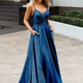 A woman stands outdoors in an elegant deep blue, floor-length gown with spaghetti straps and a v-neckline. She has her hands in the dress's pockets. A building with steps and a decorative lion figure is visible in the background, showcasing the exquisite design from MONROE PO891. Angels Formal Wear