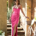 A woman in a vibrant pink, silky, spaghetti-strap KATIE PO840 stands in a sunlit, rustic archway. She is holding a white bicycle with a wicker basket. The formal KATIE PO840 features a thigh-high slit, and the background includes greenery and textured, weathered walls reminiscent of the Sunshine Coast charm. Angels Formal Wear