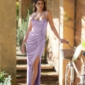 A woman in a light purple KATIE PO840 poses outdoors beside a vintage white bicycle with a woven basket. The KATIE PO840, reminiscent of formal dresses, features thin straps, a draped bodice, and a high slit. She stands under a picturesque stone archway in the sunlit garden of Sunshine Coast. Angels Formal Wear