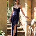 A woman in a long, elegant dark blue KATIE PO840 poses outdoors next to a vintage bicycle with a wicker basket. She stands under a columned archway with greenery in the background, capturing the charm of Sunshine Coast. The dress has a thigh-high slit and thin straps, and she rests her hand on the bicycle's handle. Angels Formal Wear