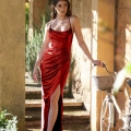 A woman in a red satin dress with thin straps and a high slit stands under a stone archway beside an old-fashioned white bicycle with a wicker basket. She gazes into the distance as she gently holds the bike's handlebar, embodying the elegance of KATIE PO840 against a rustic, Sunshine Coast backdrop. Angels Formal Wear