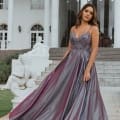 A woman stands on an outdoor staircase in front of a white building with columns, wearing one of the stunning school formal dresses—a flowing, shimmering dark silver gown with a lace bodice and thin straps. Her hair is styled in loose waves, and she looks ahead with a serene expression. Angels Formal Wear