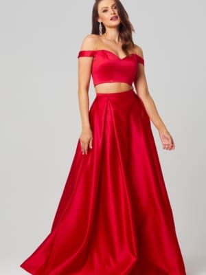 A woman stands wearing the HOLLY PO819 gown in size 12, which is a stunning, red, off-the-shoulder two-piece set featuring a fitted, midriff-baring top and a flowing skirt. She has long hair, loose earrings, and strikes a confident pose against a plain, light-colored background—ideal for browsing bridesmaids dresses in Australia or online formal wear shops. Angels Formal Wear