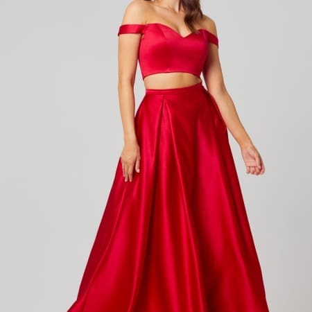 A woman stands wearing the HOLLY PO819 gown in size 12, which is a stunning, red, off-the-shoulder two-piece set featuring a fitted, midriff-baring top and a flowing skirt. She has long hair, loose earrings, and strikes a confident pose against a plain, light-colored background—ideal for browsing bridesmaids dresses in Australia or online formal wear shops. Angels Formal Wear