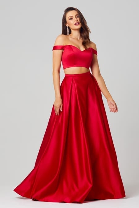 A woman stands wearing the HOLLY PO819 gown in size 12, which is a stunning, red, off-the-shoulder two-piece set featuring a fitted, midriff-baring top and a flowing skirt. She has long hair, loose earrings, and strikes a confident pose against a plain, light-colored background—ideal for browsing bridesmaids dresses in Australia or online formal wear shops. Angels Formal Wear