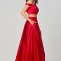 A woman with long brown hair poses in the HOLLY PO819 - Size 12, featuring a red off-the-shoulder crop top and a matching high-waisted, flowy red skirt with pockets. She rests one hand in her pocket and looks towards the camera, standing against a plain light grey background, perfect for an online formal wear shop or bridesmaids' dresses in Australia. Angels Formal Wear