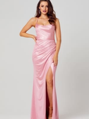 A woman stands confidently in elegant evening wear, showcasing the KATIE PO840 floor-length, sleeveless pink satin dress with thin straps and a side slit. She has one hand on her hip and is wearing high-heeled sandals. Her wavy hair is styled down, and she accessorizes with dangling earrings. Angels Formal Wear