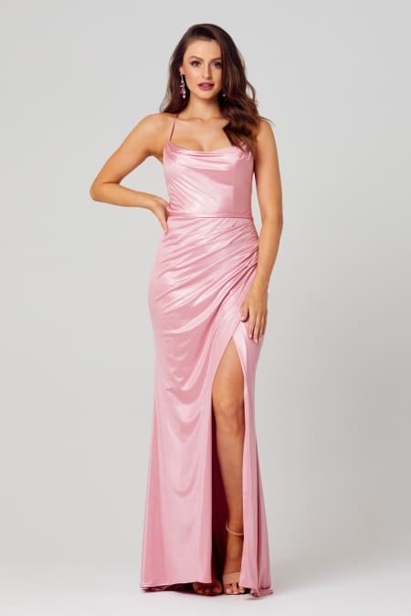 A woman stands confidently in elegant evening wear, showcasing the KATIE PO840 floor-length, sleeveless pink satin dress with thin straps and a side slit. She has one hand on her hip and is wearing high-heeled sandals. Her wavy hair is styled down, and she accessorizes with dangling earrings. Angels Formal Wear