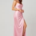 A woman is standing and wearing the KATIE PO840, a pink satin dress with spaghetti straps and a high slit on one side, perfect for evening wear. She has her arms relaxed by her sides and is looking slightly to her left. The background is light gray. Angels Formal Wear