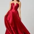 A woman wearing a strapless, red satin evening gown poses against a plain background. The dress, resembling designer bridesmaid dresses, features a sweetheart neckline and a flowing, floor-length skirt. She is holding one side of the skirt, creating a graceful flare. She has long, wavy hair and is wearing drop earrings. Angels Formal Wear