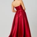 A woman with long dark hair stands with her back to the viewer, wearing a strapless, floor-length, red satin gown from a designer bridesmaid dresses collection. She also wears large, dangling earrings. The background is plain and gray. Angels Formal Wear