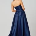 A woman with long, dark hair stands with her back to the camera, wearing a strapless, floor-length navy blue gown. The fabric of this designer bridesmaid dress is shiny and smooth, cascading elegantly to the ground. She accessorizes with long, drop earrings. Angels Formal Wear