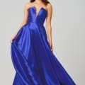 A woman poses in a vibrant blue, floor-length, sleeveless gown with a plunging neckline. She has long, wavy hair and is wearing earrings. She holds the sides of her designer bridesmaid dress, creating a flowing effect. The background is plain and light-colored. Angels Formal Wear