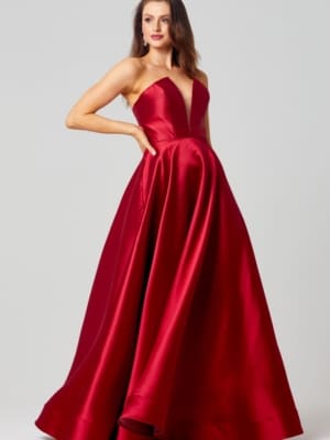 A woman wearing a strapless, floor-length, red satin gown with a fitted bodice and full skirt poses with one hand on her hip. She has long, wavy hair and is standing against a plain, light gray background. The elegant gown could be a highlight in the realm of formal dresses or designer bridesmaid dresses. Angels Formal Wear