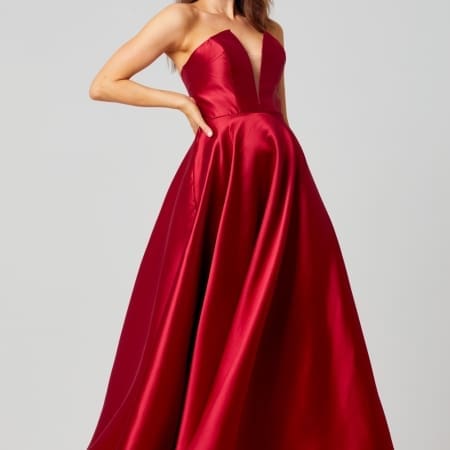 A woman wearing a strapless, floor-length, red satin gown with a fitted bodice and full skirt poses with one hand on her hip. She has long, wavy hair and is standing against a plain, light gray background. The elegant gown could be a highlight in the realm of formal dresses or designer bridesmaid dresses. Angels Formal Wear