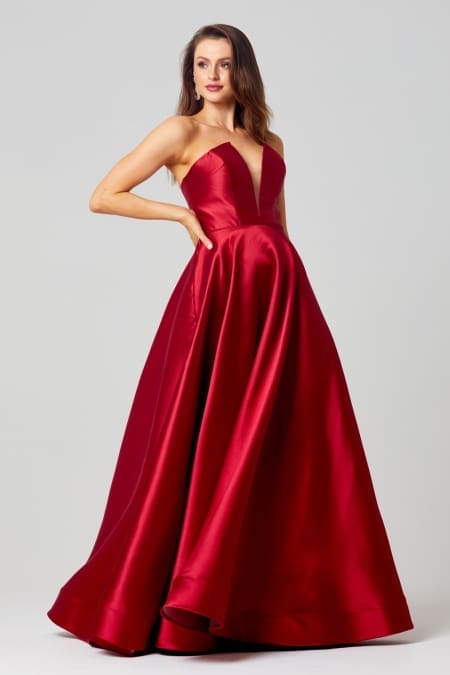 A woman wearing a strapless, floor-length, red satin gown with a fitted bodice and full skirt poses with one hand on her hip. She has long, wavy hair and is standing against a plain, light gray background. The elegant gown could be a highlight in the realm of formal dresses or designer bridesmaid dresses. Angels Formal Wear