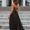 A woman stands on a flight of stairs leading to a large building with glass doors. She is wearing one of the stunning school formal dresses, a long, black gown with an intricate lace-up back design. Her hair is styled in loose waves, and she is looking over her left shoulder. Angels Formal Wear