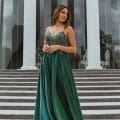 A woman stands confidently on a set of stone steps in front of a building with tall columns, wearing one of the stunning school formal dresses—a long, flowing green dress with intricate detailing on the bodice and straps. She gazes off into the distance with a contemplative expression. Angels Formal Wear