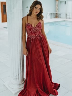 A woman in a luxurious red gown stands next to a white pillar. The dress features intricate lace detailing on the bodice and thin straps, exemplifying stunning school formal dresses. She is in an elegant indoor setting with a pristine pool in the background. The overall ambiance is sophisticated and serene. Angels Formal Wear