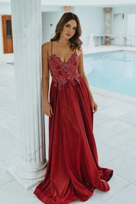 A woman in a luxurious red gown stands next to a white pillar. The dress features intricate lace detailing on the bodice and thin straps, exemplifying stunning school formal dresses. She is in an elegant indoor setting with a pristine pool in the background. The overall ambiance is sophisticated and serene. Angels Formal Wear