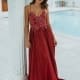 A woman in a luxurious red gown stands next to a white pillar. The dress features intricate lace detailing on the bodice and thin straps, exemplifying stunning school formal dresses. She is in an elegant indoor setting with a pristine pool in the background. The overall ambiance is sophisticated and serene. Angels Formal Wear