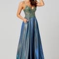 A woman poses in a shimmering, sleeveless blue gown with a fitted, embellished bodice and flowing skirt. She stands against a plain, light gray backdrop, touching her hair with a relaxed smile and her other hand resting in a pocket of the designer bridesmaid dress. Angels Formal Wear