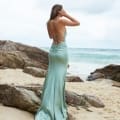 A woman stands on a rocky beach facing the ocean, wearing one of the stunning school formal dresses—a long, light teal gown with a low back and thin straps. Her hand is raised to her head, and her hair hangs loose. The sky is overcast, and waves are gently crashing in the background. Angels Formal Wear