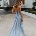 A woman with wavy hair is wearing a MONROE PO891, a light blue backless evening gown with crisscross straps, standing outdoors on a paved area. The Sunshine Coast structure behind her features white walls, large windows, and ornate statues of lions. Angels Formal Wear